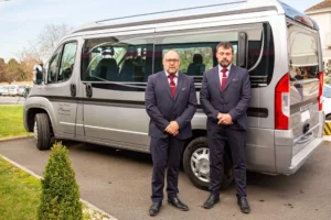 Deux porteurs funéraires en costume posant devant un véhicule de transport gris.